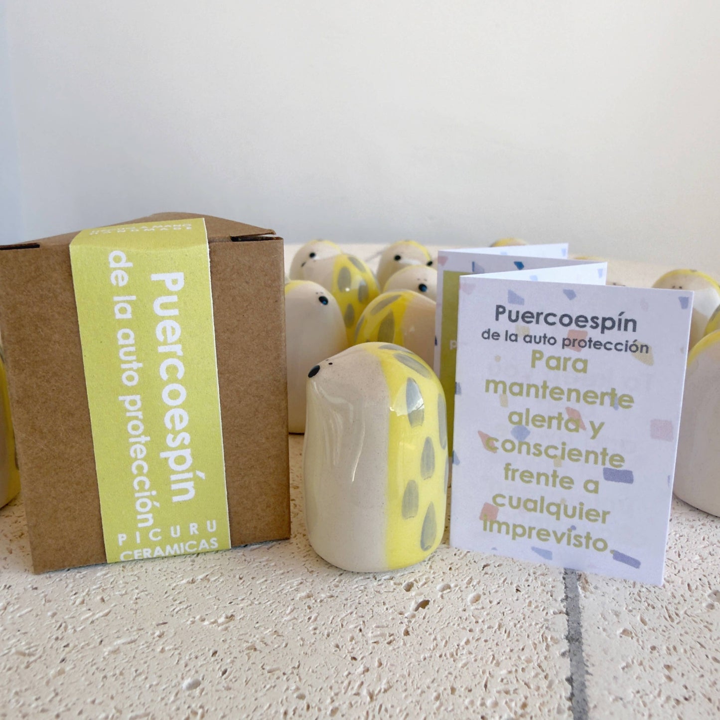 The Porcupine Amulet of Self-Protection by Picuru ceramicas, a handmade ceramic figurine symbolizing awareness and strength, stands by a brown box and an informational card in Spanish. Additional amulets are seen in the background.
