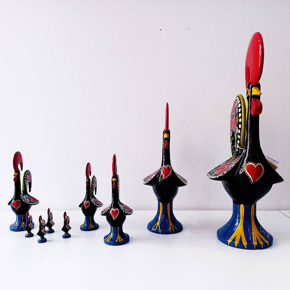 A lineup of Tejo Shop Barcelos Rooster figurines, decorated with red crests, heart motifs, and detailed wings, is displayed in size order on a white surface against a plain white background.