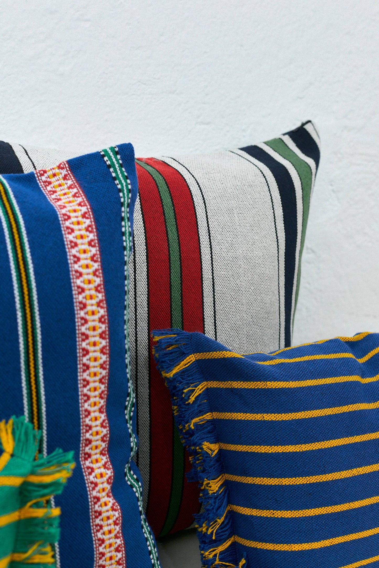 Three vibrant woven pillows, including the Blue Pomegranate Cushion Cover 50cmx45cm by Eturel Madrid, handmade in Spain with blue, yellow, and white stripes plus fringe, displayed on a crisp white background.