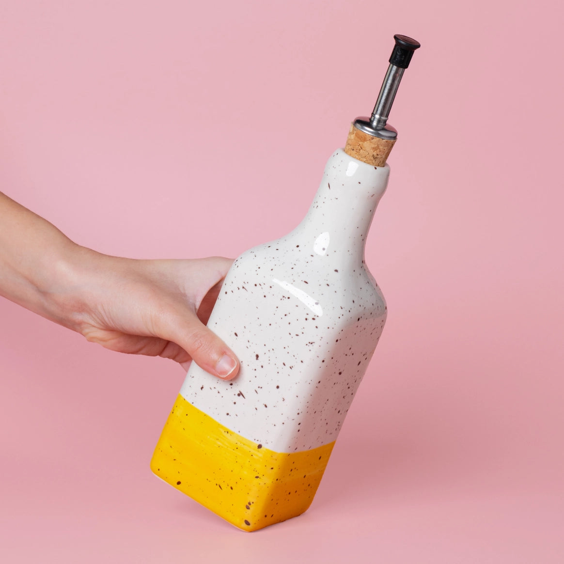 A hand holds the CANELA Oil and Salad Bowl from Alfar Tierra Cocida—a speckled white ceramic oil dispenser with yellow base and metal pour spout—part of a handmade kitchenware set, shown against a pink background.