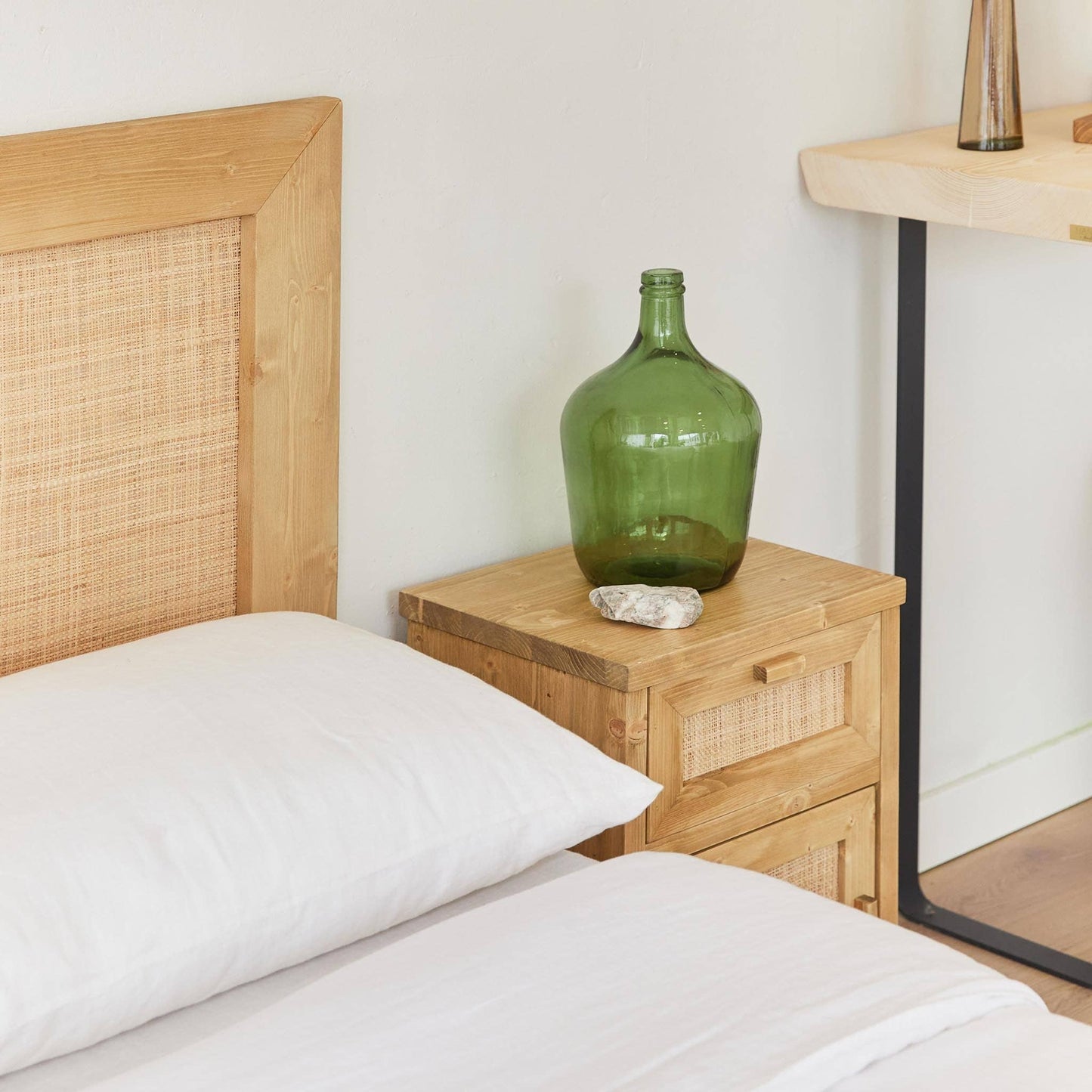 A neatly made bed with white linens sits beside a nightstand featuring the Luiza Demijohn Vase by Hannun, crafted sustainably. A wooden desk with a black leg appears in the background.