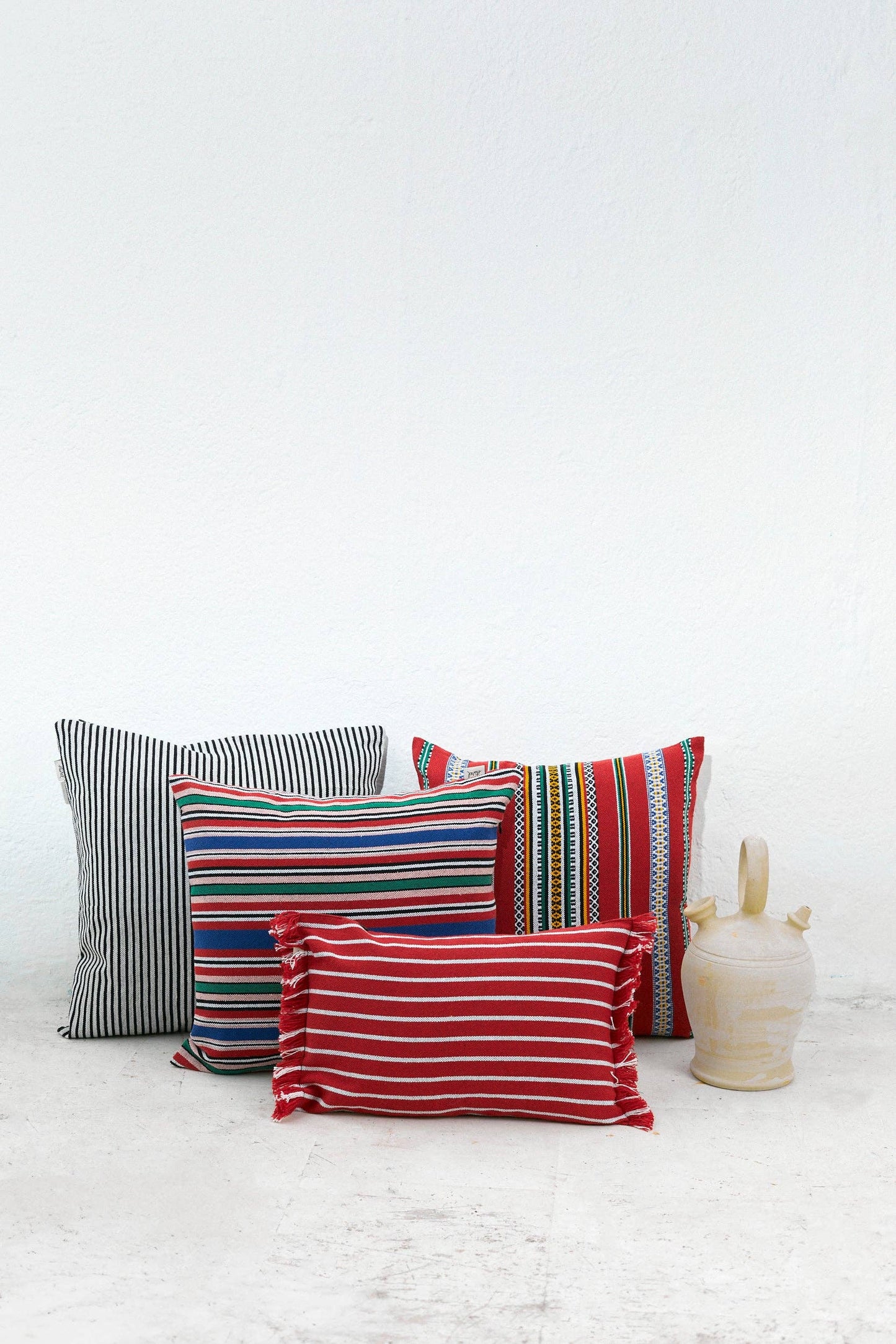 Four Eturel Madrid Red Pomegranate Cushion Covers (50cm x 45cm) with black, white, red, yellow, and blue stripes are arranged by a white wall near a light ceramic jug. Each cover is handmade and inspired by Spanish craftsmanship.