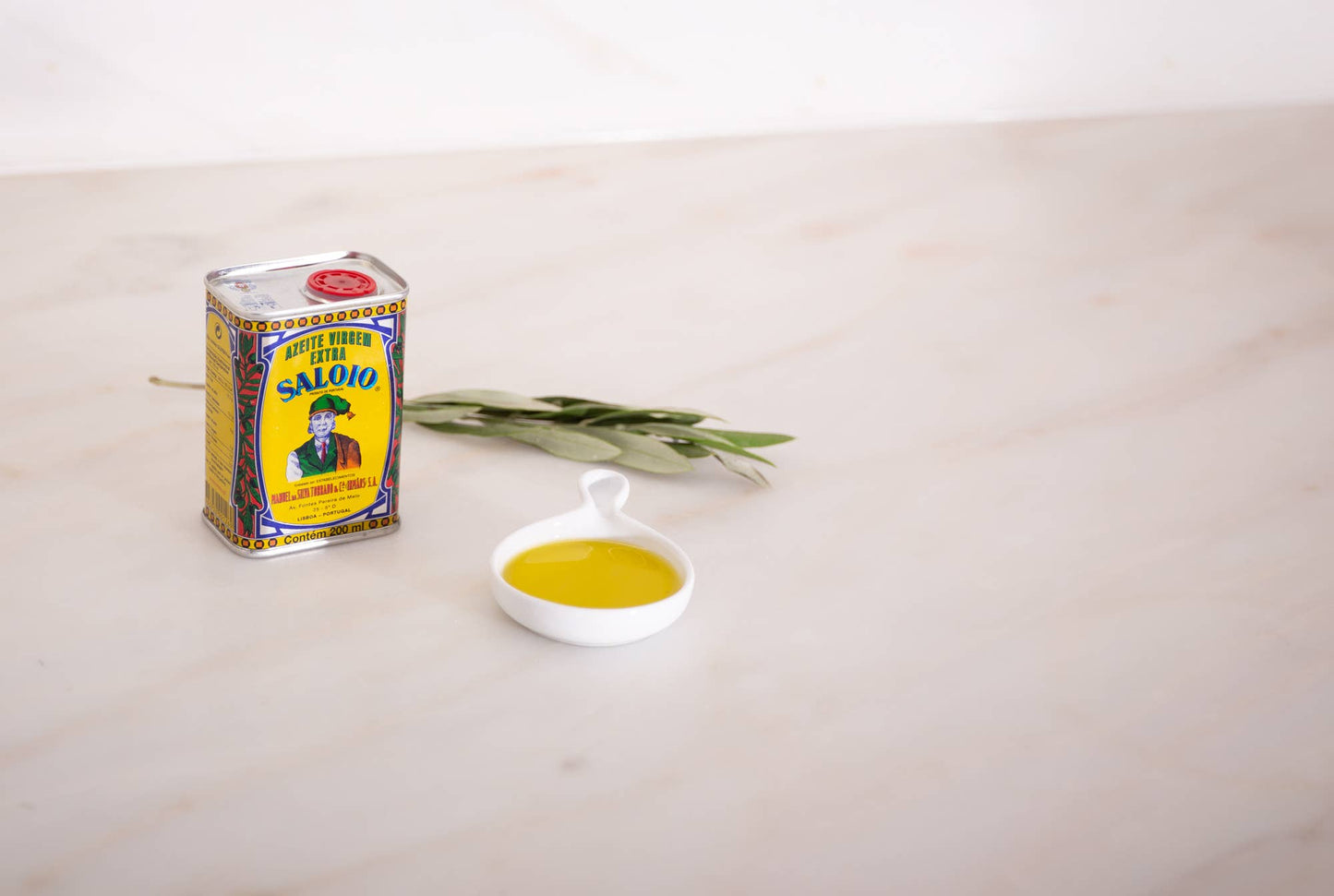 A 200ml can of Machado’s Saloio Olive Oil from Portugal sits beside a white dish with olive oil and olive leaves, showcasing Portuguese tradition on a light marble surface.