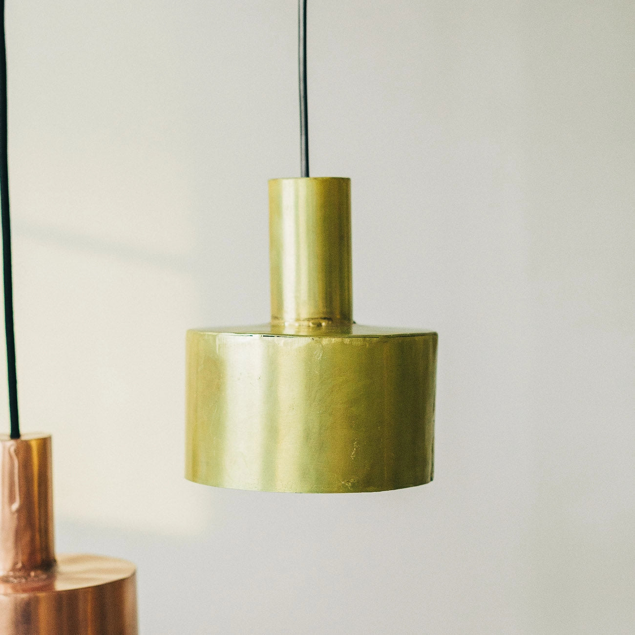 The Seber Lamp by Hannun, shown here in brass with a cylindrical design and black cord, is perfect for industrial-inspired interiors. A copper version of the Seber Ceiling Lamp is partially visible on the left. Available in 3 colours.
