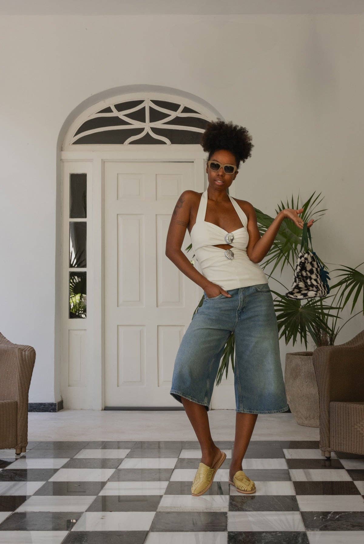 A woman stands on a checkered floor, wearing sunglasses, a white halter top, long denim shorts, and Mexas EU’s Yellow San Pancho Slide Mules with a cushioned footbed. She holds a checkered bag between wicker chairs and potted plants.