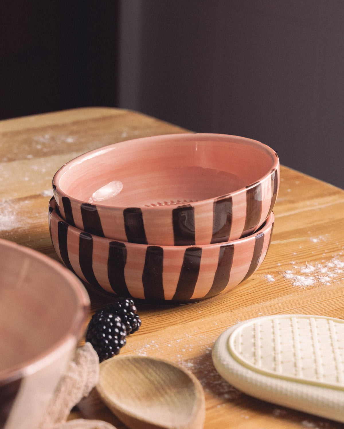 Two stacked ceramic bowls with pink and dark brown stripes sit on a wooden table, next to a wooden spoon, pot holder, some flour, a blackberry, and the Alfar Tierra Cocida Large Pottery Grater Plate – Chocolate NEW.
