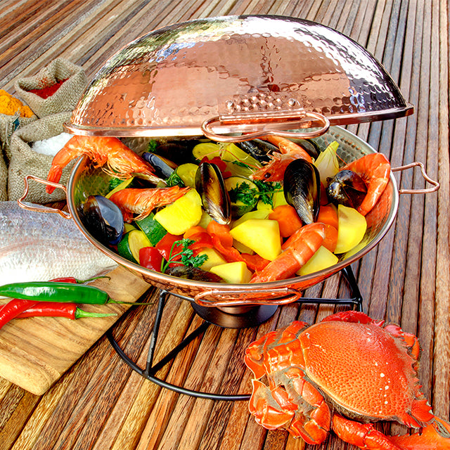 The Cataplana starter kit by Iberica - Pretty things from Portugal sits on a wooden table, its copper pot brimming with shrimp, mussels, potatoes and herbs. A whole crab, chili peppers and raw fish beside it evoke authentic Portuguese cuisine.