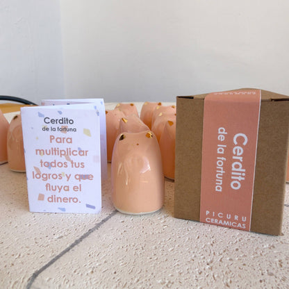 The Fortune Pig Amulet - Gold Luster by Picuru ceramicas sits between a Spanish card and a brown "Cerdito de la fortuna" box, with more golden pig figurines in the background on a cream surface.