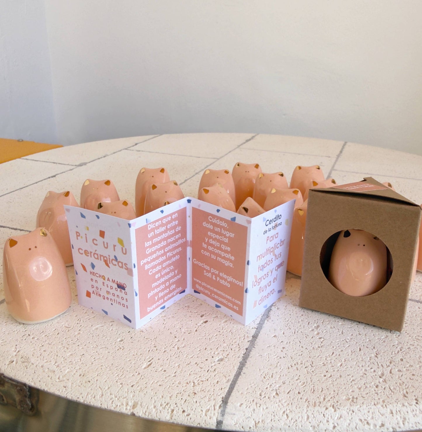 A display of Picuru ceramicas’ Fortune Pig Amulet - Gold Luster 5 Languages shows small pink piggy figurines on white tiles, with folded info cards and a boxed amulet featuring a cutout window in front.
