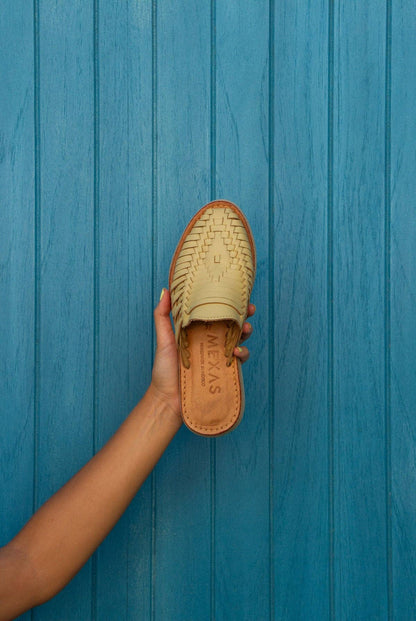 A hand holds a Yellow San Pancho Slide Mule by Mexas EU, featuring intricate woven leather and a cushioned footbed, set against a blue wooden background.