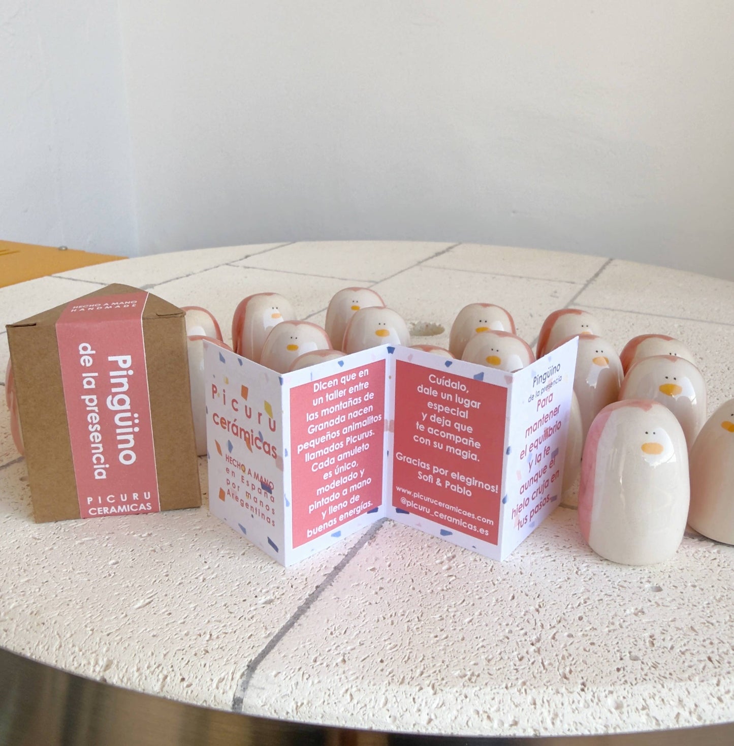 A set of handmade Penguin Amulet of Presence figurines by Picuru ceramicas, symbols of balance and resilience, are arranged beside a brown box labeled with the product name and an open pamphlet featuring red and white text.