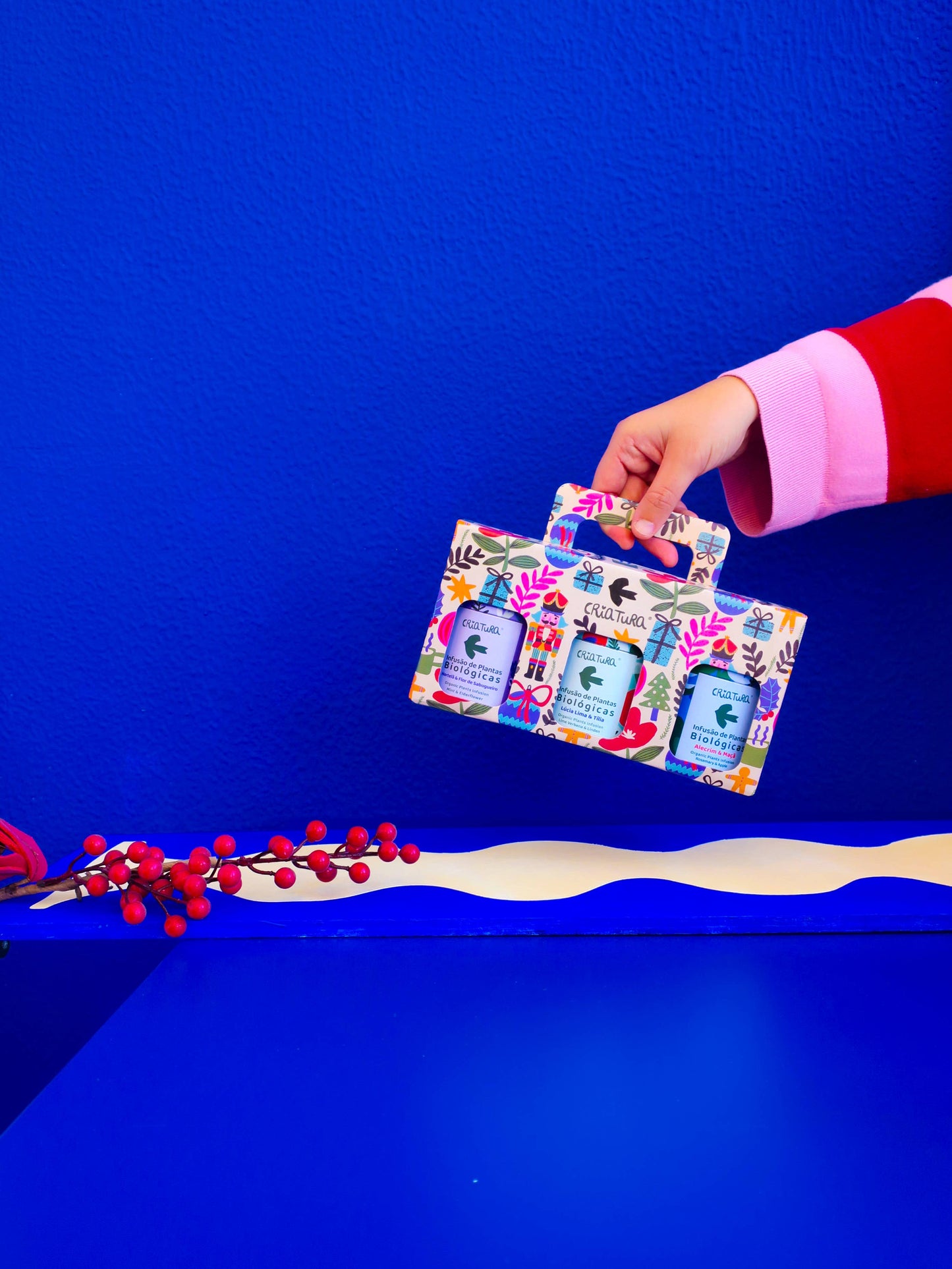 A hand in a red and pink sleeve holds Criatura’s Christmas Tea Box | Criatura X Teresa Rego - Special Edition with three bath products, set against a vibrant blue background and red berries in the lower left corner.