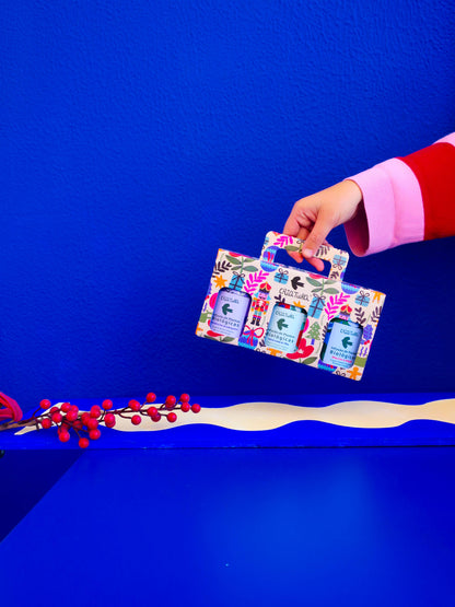 A hand in a red and pink sleeve holds Criatura’s Christmas Tea Box | Criatura X Teresa Rego - Special Edition with three bath products, set against a vibrant blue background and red berries in the lower left corner.