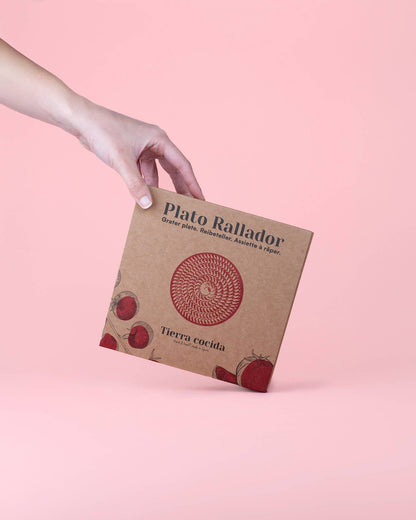 A hand holds a brown box labeled Alfar Tierra Cocida, with a red circle and tomato images. Inside is the Garlic Grater Plate - Medium Ceramic - Azul, a hand-crafted Spanish kitchen tool, set against a solid light pink background.