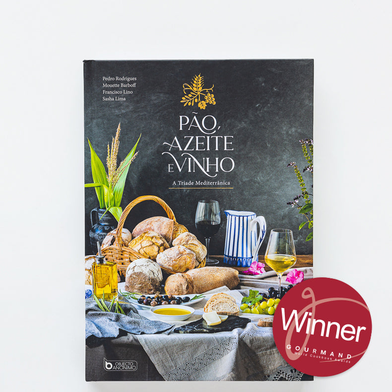 The cookbook "Bread, Olive Oil & Wine" by ObjectoAnonimo stands upright amid Mediterranean staples—bread, cheese, wine, and olive oil. A red Winner Gourmand badge is visible in the lower right corner.