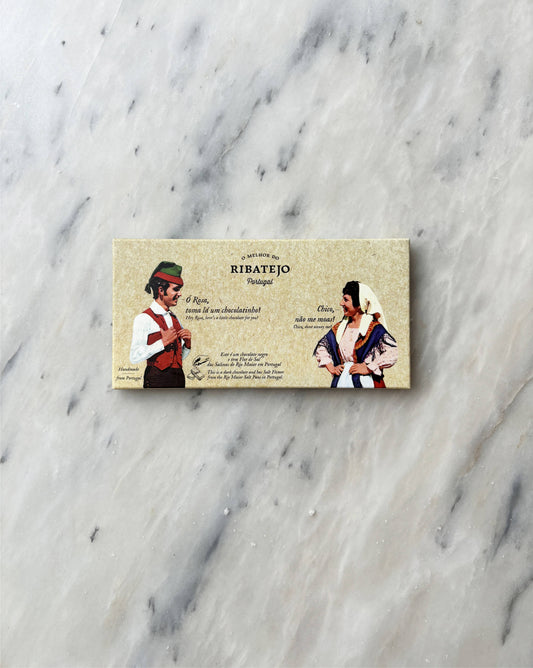 A rectangular package of O Melhor do Ribatejo Dark Chocolate with Flor de Sal, featuring a man and woman in traditional Portuguese clothing and Portuguese text on a beige background, rests on a white marble surface.