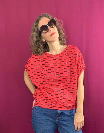 A woman with curly hair poses before a magenta background, wearing oversized dark sunglasses, blue jeans, and La Compiano’s Lola T-Shirt with a sausage print, made in Spain. She stands confidently, hand on hip and head tilted.