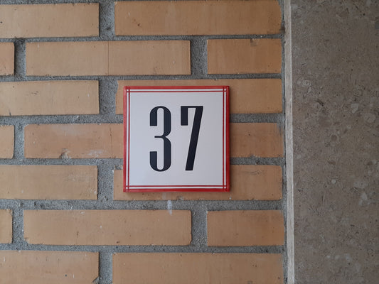 A white square TilePassion Handpainted Ceramic Door Number (37) in bold black font with a red border, styled like custom Portuguese tiles, is mounted on a yellow brick wall beside gray stone.
