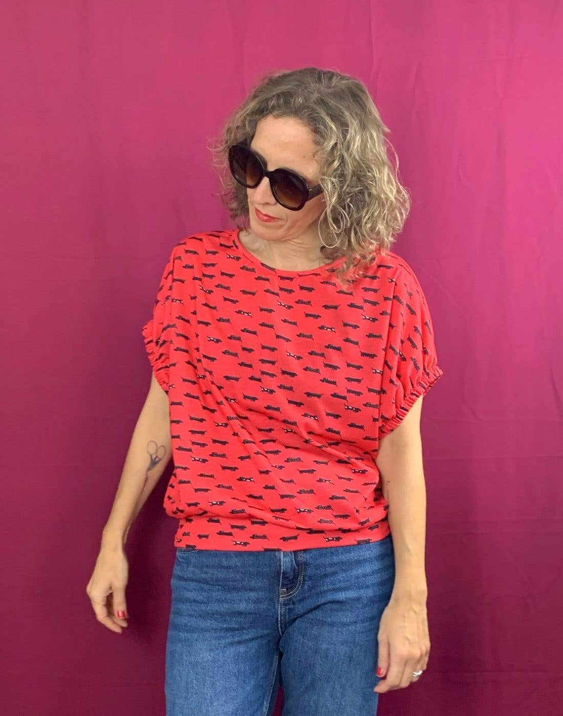 A woman with curly blonde hair and large dark sunglasses wears the La Compiano Lola T-Shirt - Sausage Print and blue jeans, standing against a bright pink background while looking slightly down and to the side.