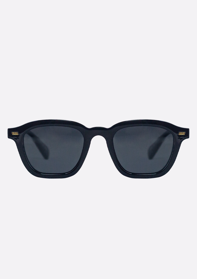 The Oceanides Neda Shiny Black Sunglasses feature a recycled plastic frame, dark polarized lenses, and gold corner accents, displayed against a plain light background.