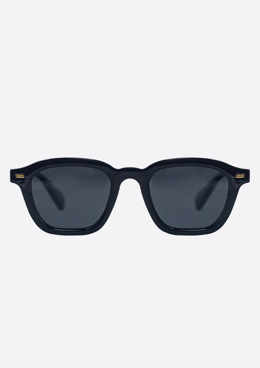The Oceanides Neda Shiny Black Sunglasses feature a recycled plastic frame, dark polarized lenses, and gold corner accents, displayed against a plain light background.