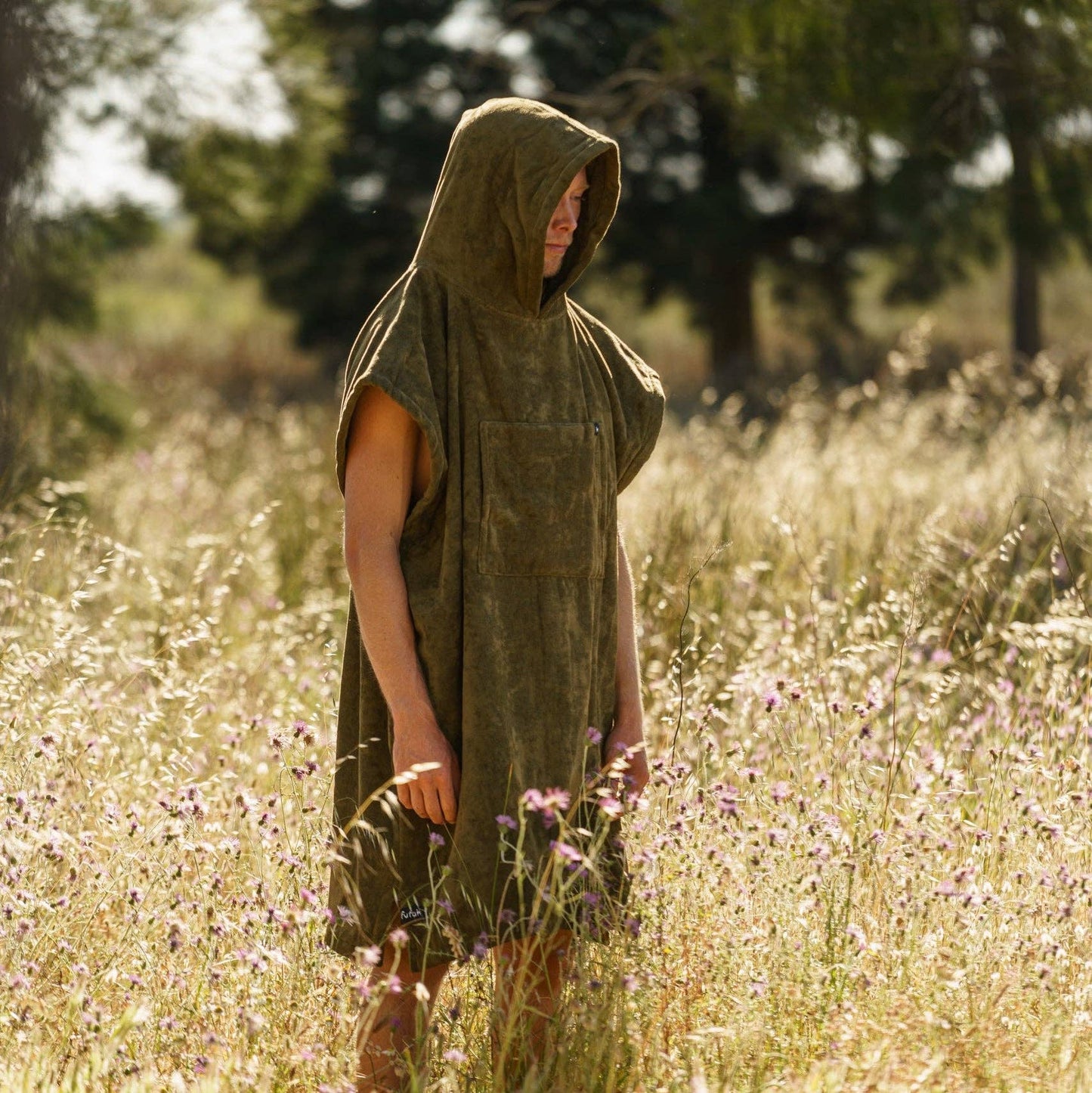 A person wearing the Futah Ericeira Terry Poncho, a sleeveless olive-green cotton hooded piece, stands in a sunlit field of tall grass and wildflowers with blurred trees in the background.