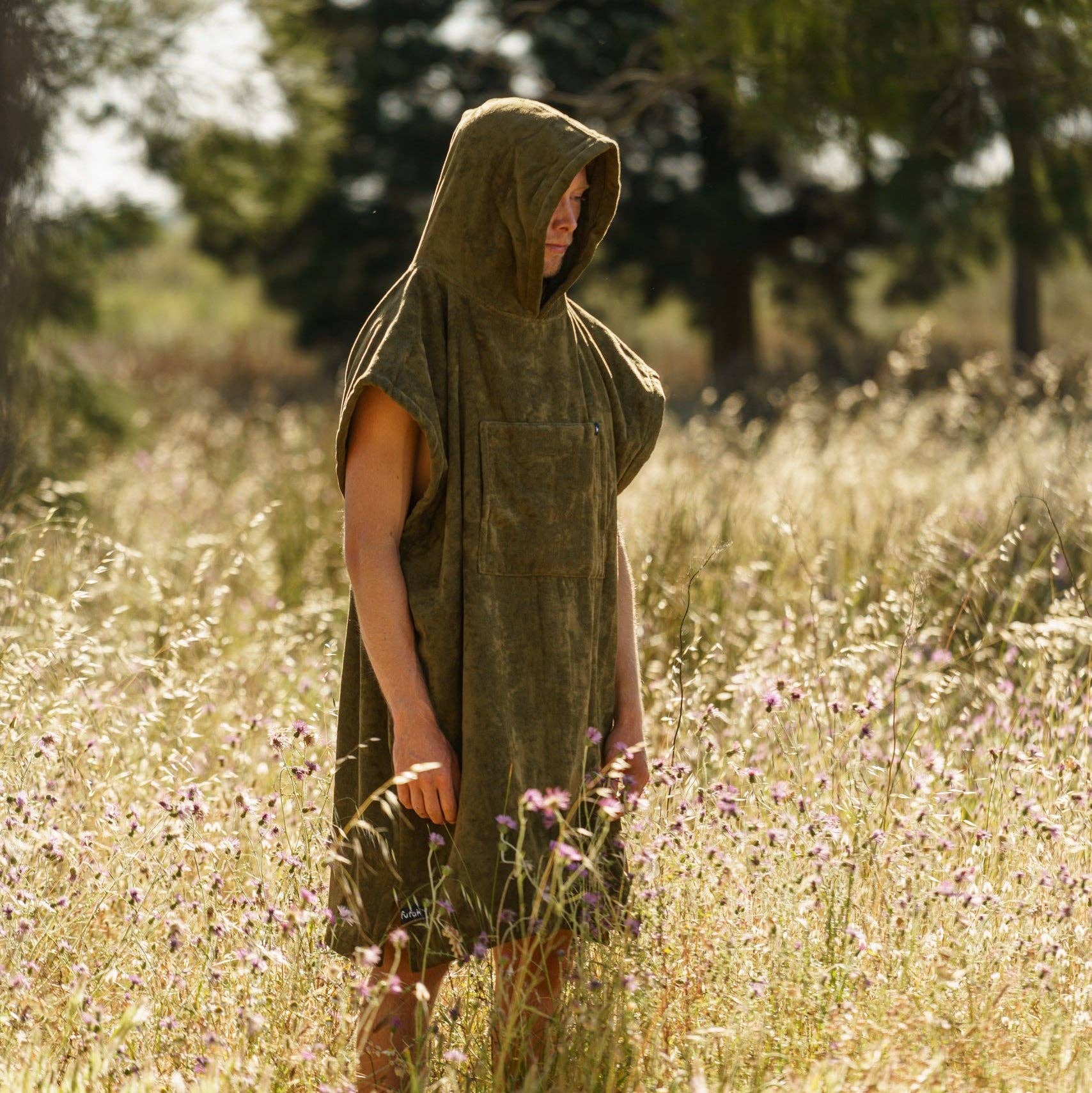 A person wearing the Futah Ericeira Terry Poncho, a sleeveless olive-green cotton hooded piece, stands in a sunlit field of tall grass and wildflowers with blurred trees in the background.