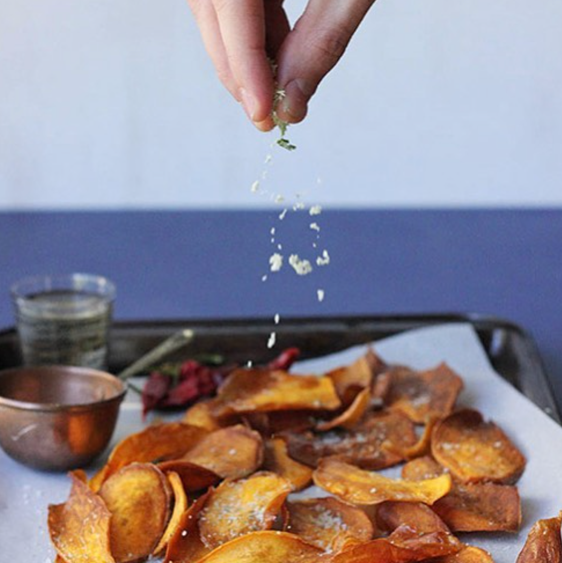A hand sprinkles O Melhor do Ribatejo's Salt Flower | From Portugal over golden-brown potato chips on a baking sheet, evoking authentic Ribatejo flavor, with a small cup and dish of seasoning nearby.