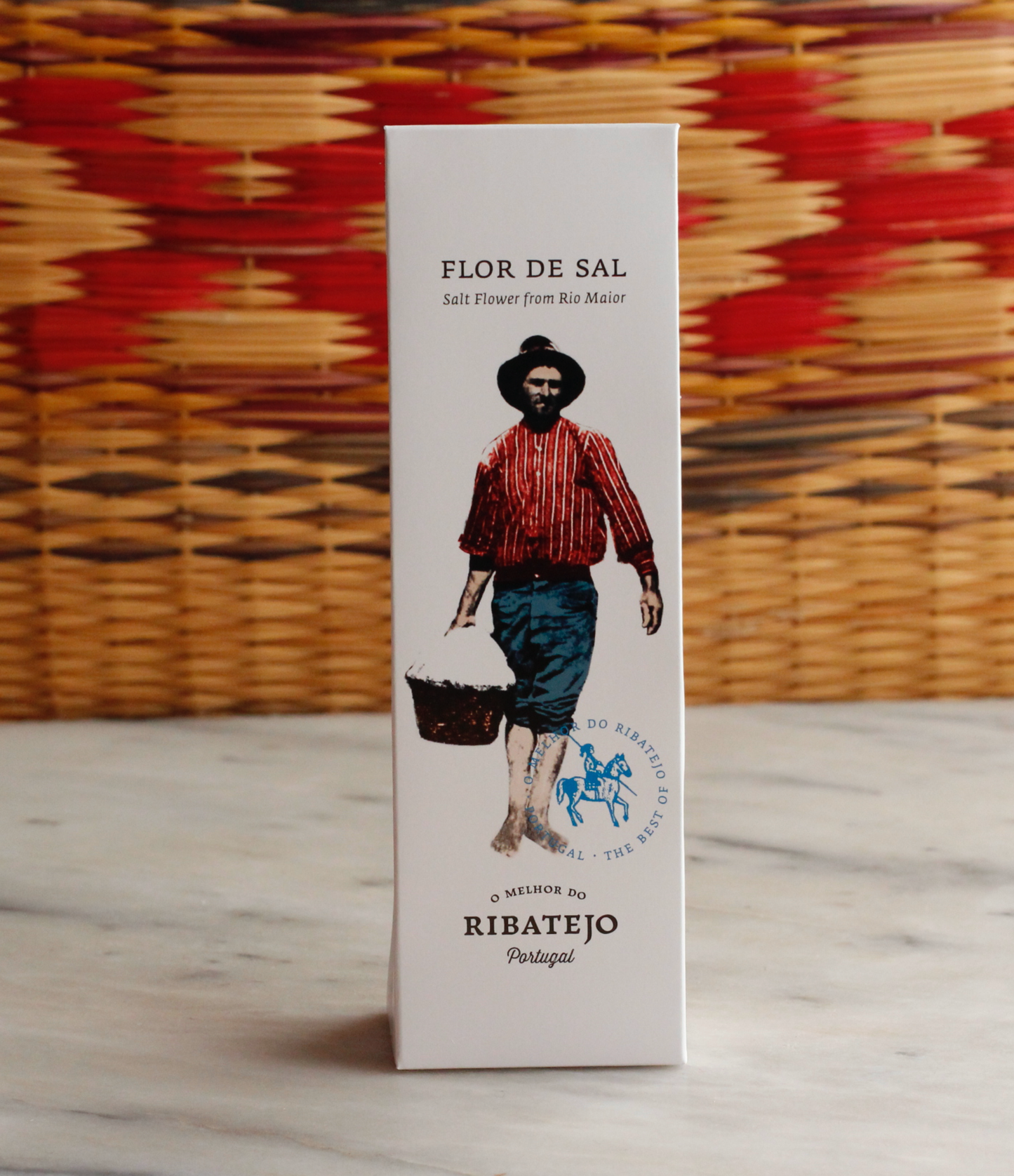 A white box of O Melhor do Ribatejo Salt Flower | From Portugal, featuring an illustration of a person in traditional clothing with a basket, set against a woven basket background.