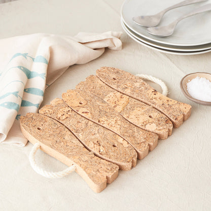 The LIGA Natural Cork Trivet - Fish features an eco-friendly cork design inspired by Portugal, with white rope handles. It's displayed on a beige tablecloth near a folded napkin, plates, spoons, and a small bowl of salt.
