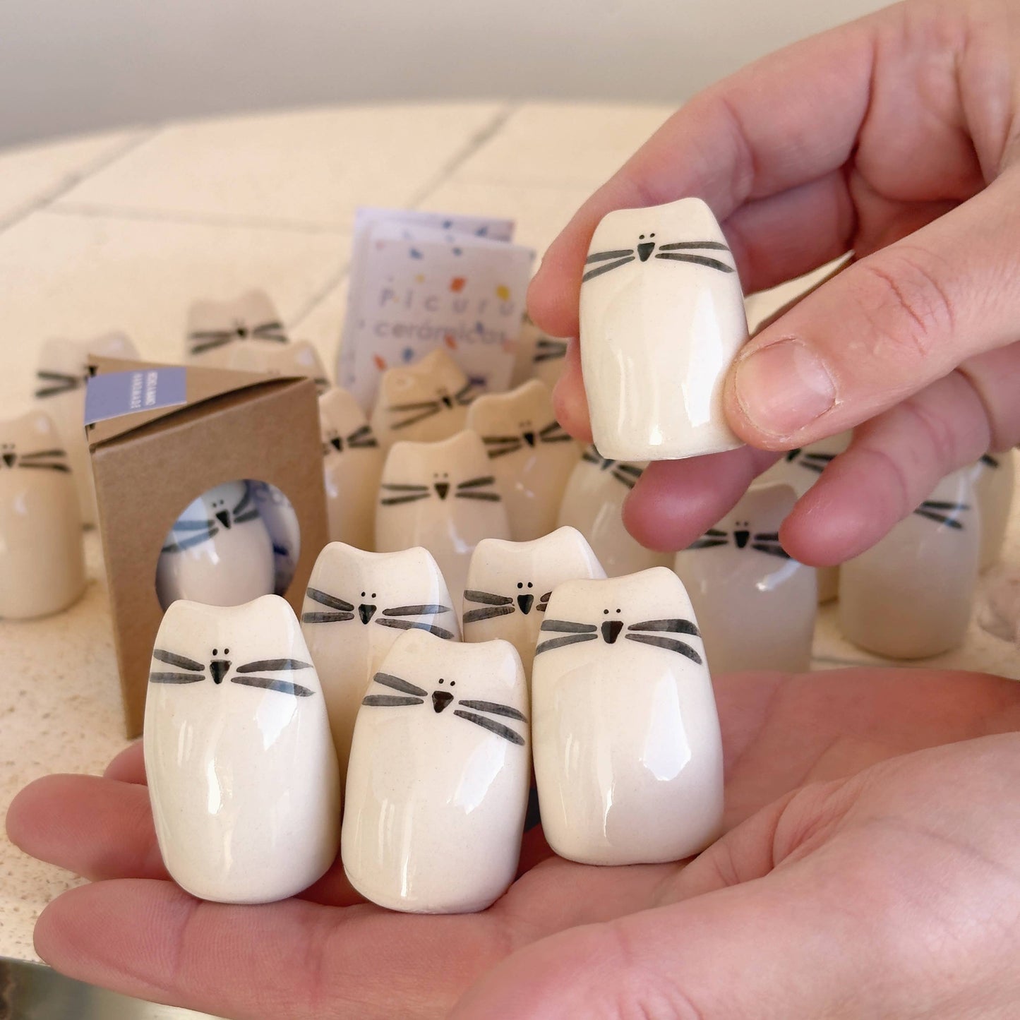 A person holds several Kitten Amulet of Cuddles—handmade ceramic cat figurines by Picuru ceramicas, each a thoughtful friendship and comfort gift. More minimalist kitten amulets are arranged on the table in the background.
