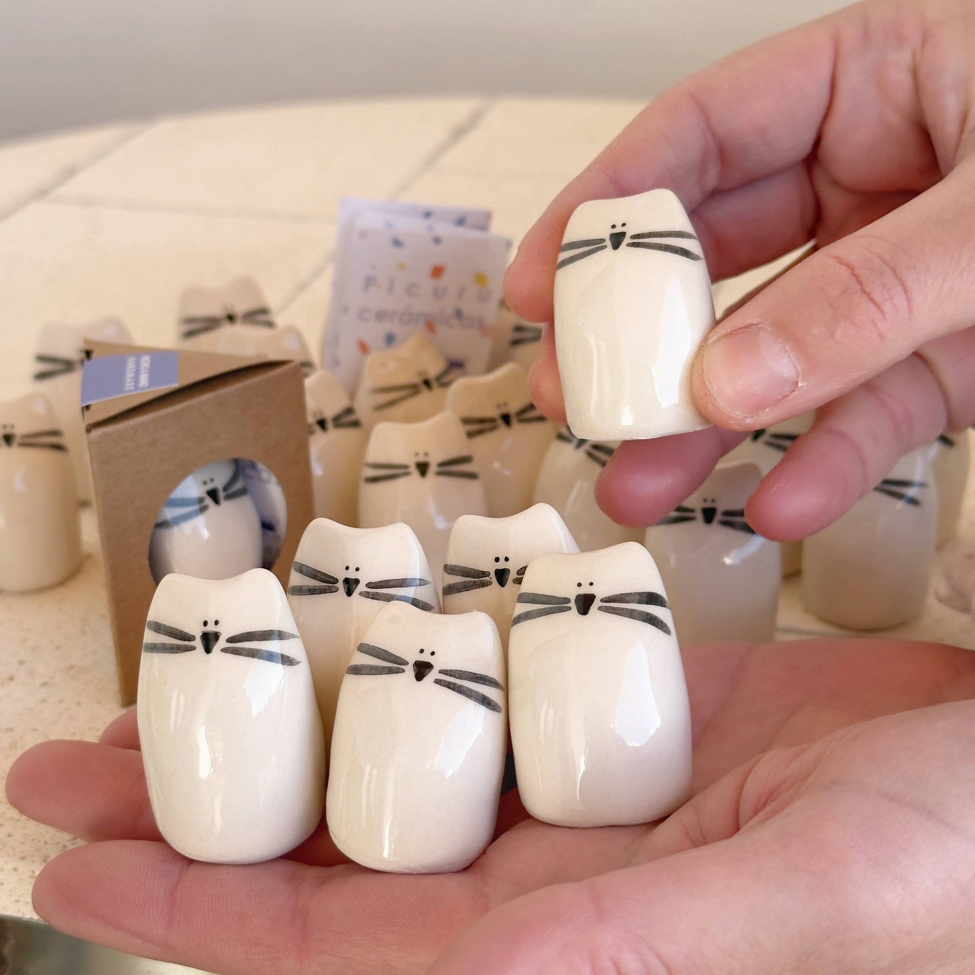 A person holds several Kitten Amulet of Cuddles—handmade ceramic cat figurines by Picuru ceramicas, each a thoughtful friendship and comfort gift. More minimalist kitten amulets are arranged on the table in the background.
