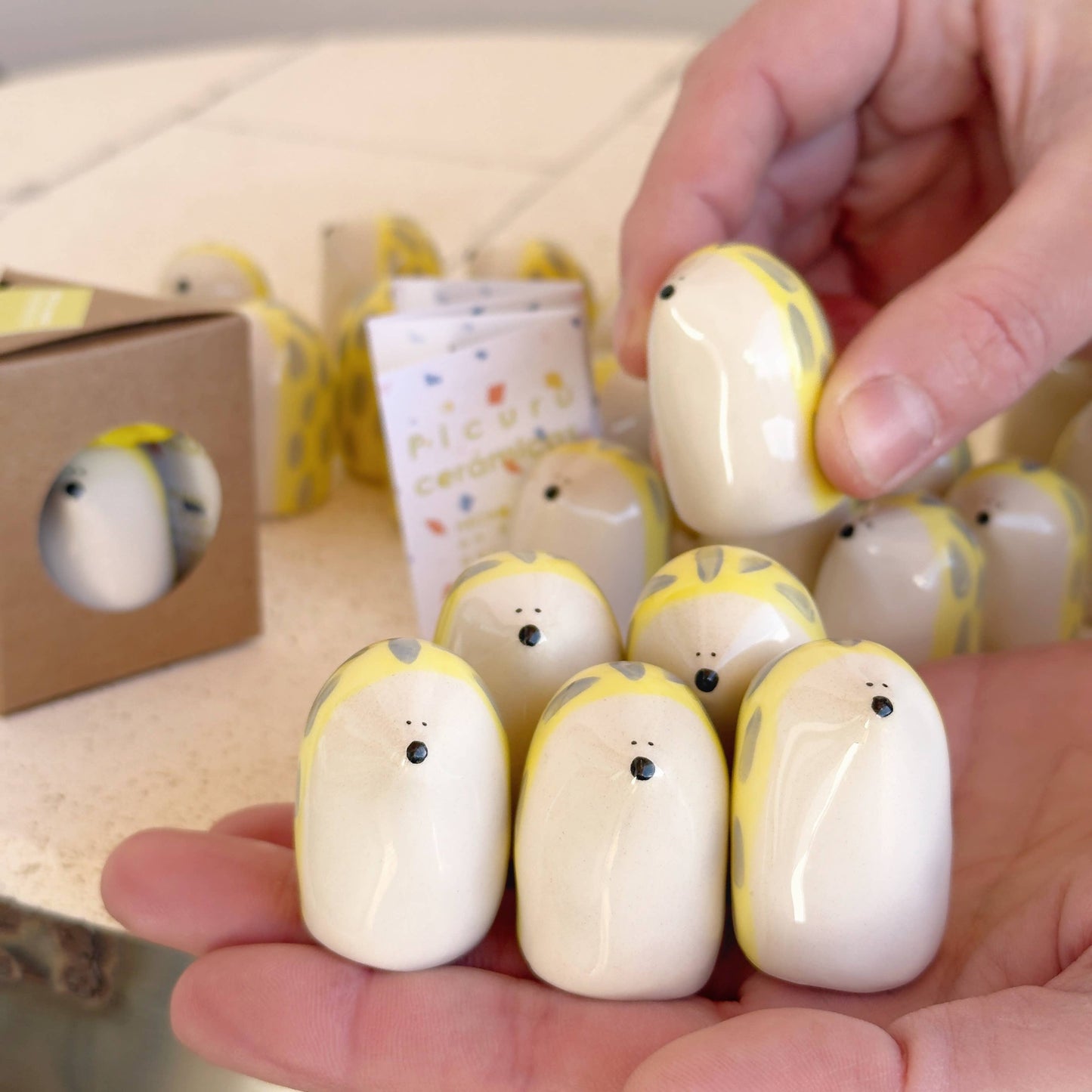 A person holds several Picuru ceramicas’ Porcupine Amulet of Self-Protection—handmade ceramic figures with yellow spots and tiny black eyes and noses. More amulets and a cardboard box appear on the table in the background.