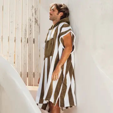A person stands against a white wall, wearing the Bedu Terry Poncho by Futah, featuring wide vertical brown and white stripes. Sunlight casts soft shadows on the Portuguese cotton poncho, creating a relaxed, summery atmosphere.
