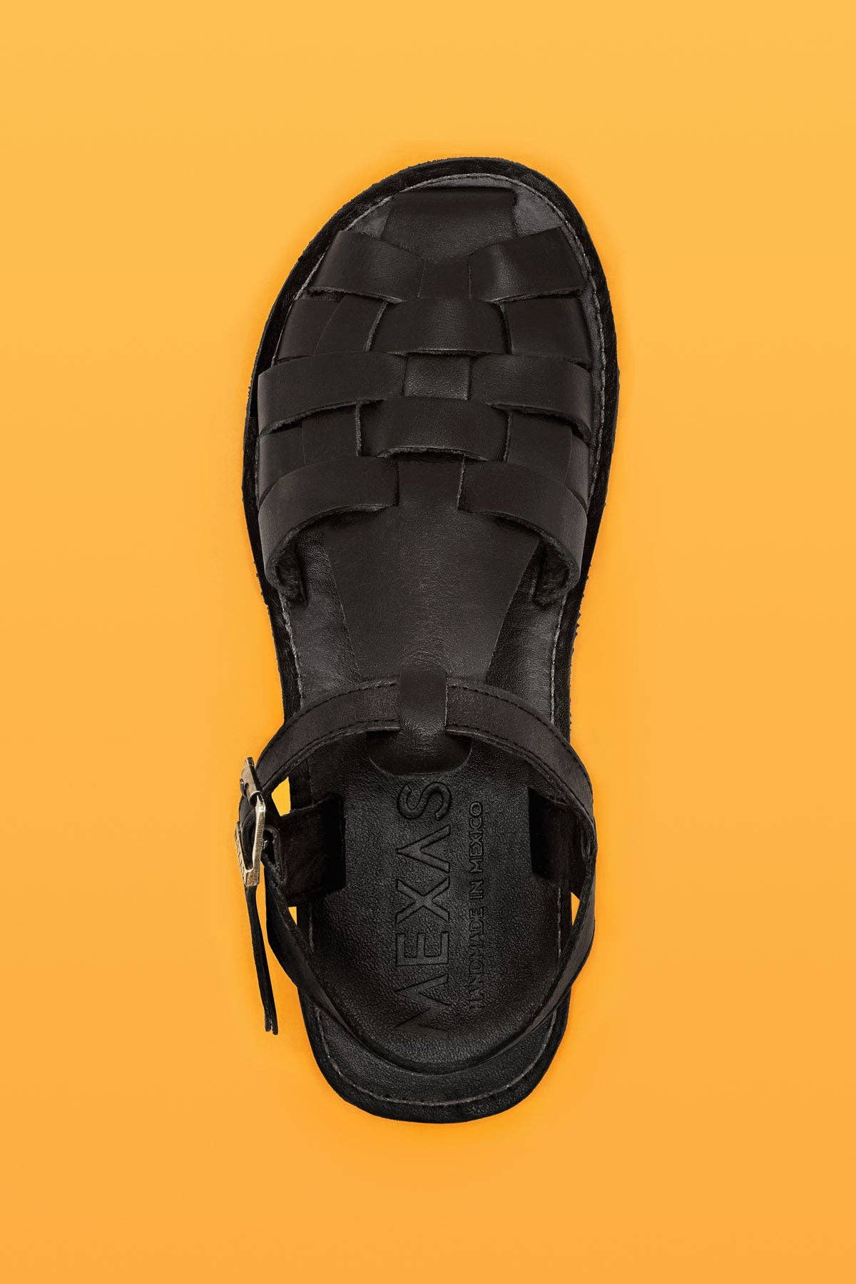 A single Mexas EU Black Glory sandal, made in Spain with woven leather and a buckle strap, is shown from above on a solid orange background.