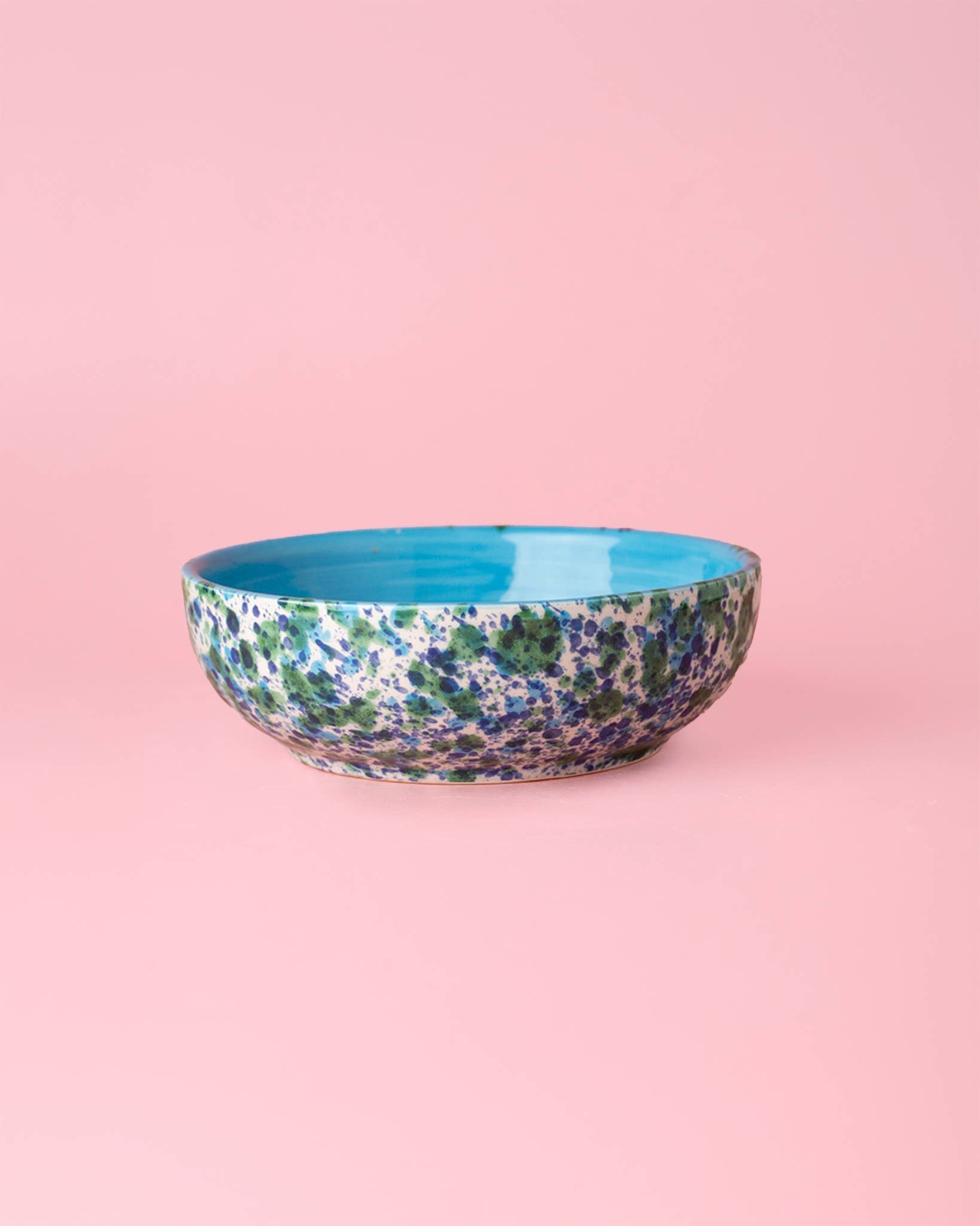 The Alfar Tierra Cocida Garlic Grater Plate - Medium Ceramic - Azul features a blue, green, and purple speckled exterior with a solid turquoise interior, displayed on a pink background.