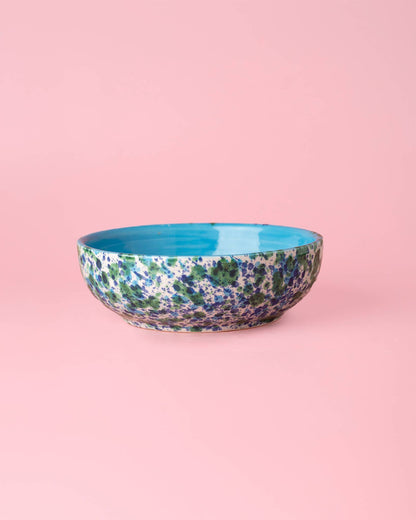 The Alfar Tierra Cocida Garlic Grater Plate - Medium Ceramic - Azul features a blue, green, and purple speckled exterior with a solid turquoise interior, displayed on a pink background.