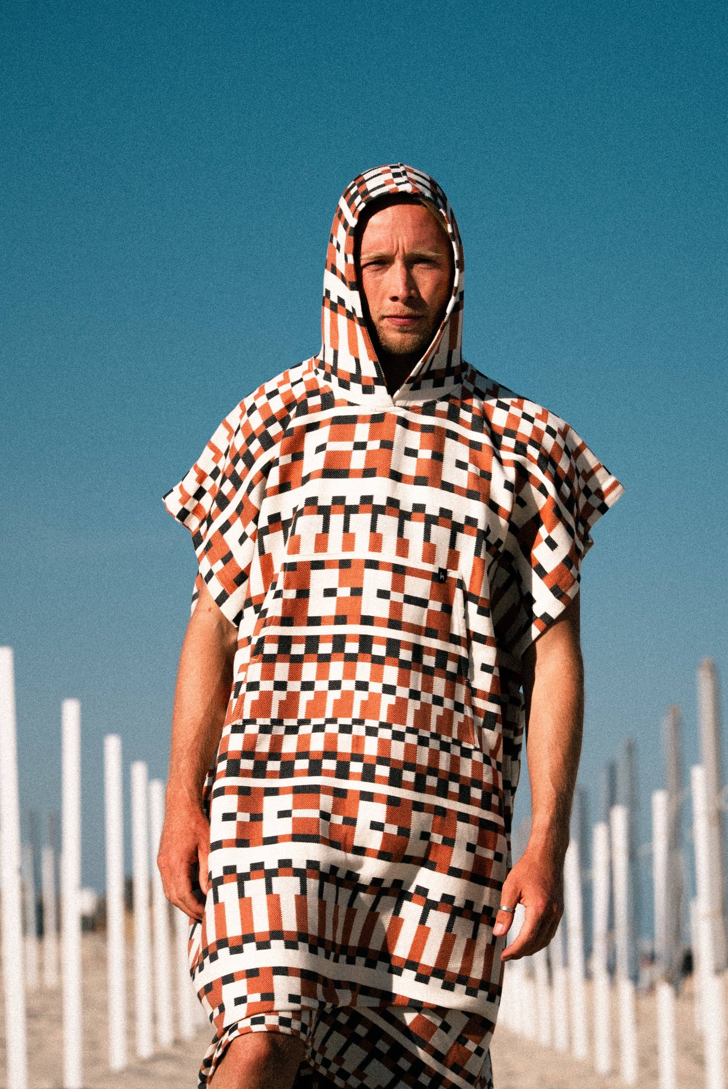 A person stands on a beach wearing the Futah Cacela Poncho, a bold and lightweight cotton piece featuring striking black, white, and orange geometric patterns. Against a clear blue sky with white poles in the background, it's the perfect attire for enjoying Portuguese beachwear in style.