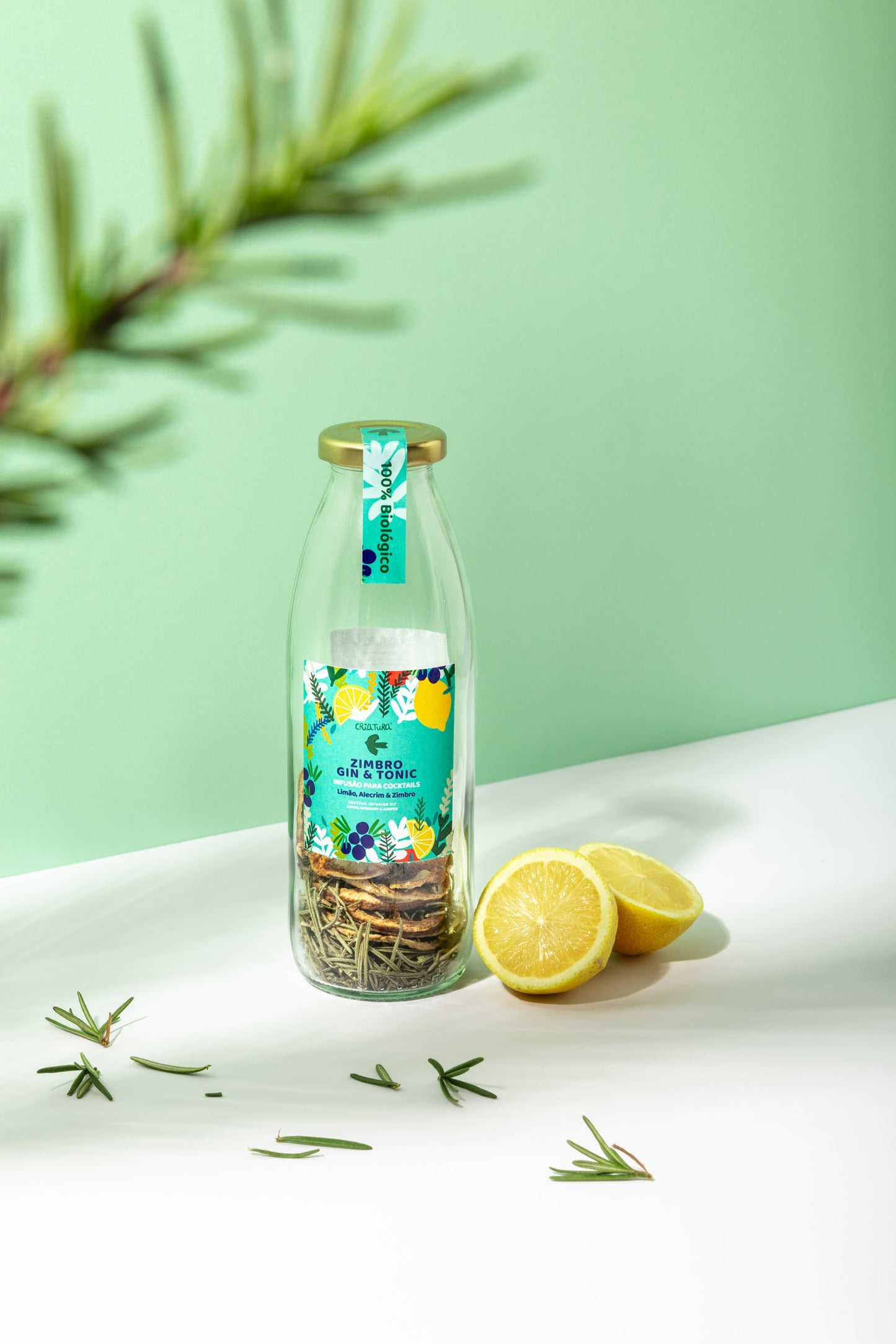 The Criatura Organic Cocktail Infusion – Lemon, Fennel & Juniper sits on a white surface with a halved lemon and rosemary sprigs, perfect for home mixology and displayed against a green background.