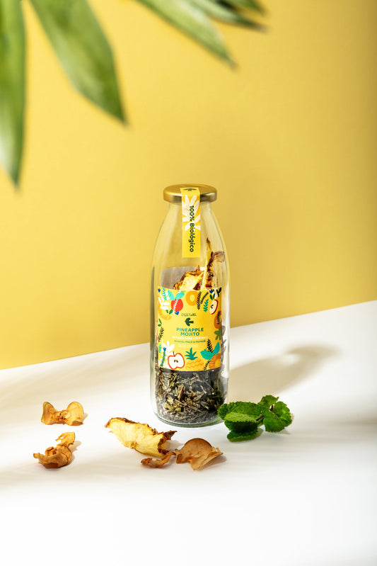 A glass bottle of Criatura’s Organic Mojito Cocktail Infusion – Pineapple, Apple & Mint sits on a white surface, surrounded by dried fruit and green leaves. Its colorful label pops against a yellow background—ideal for craft cocktails at home.