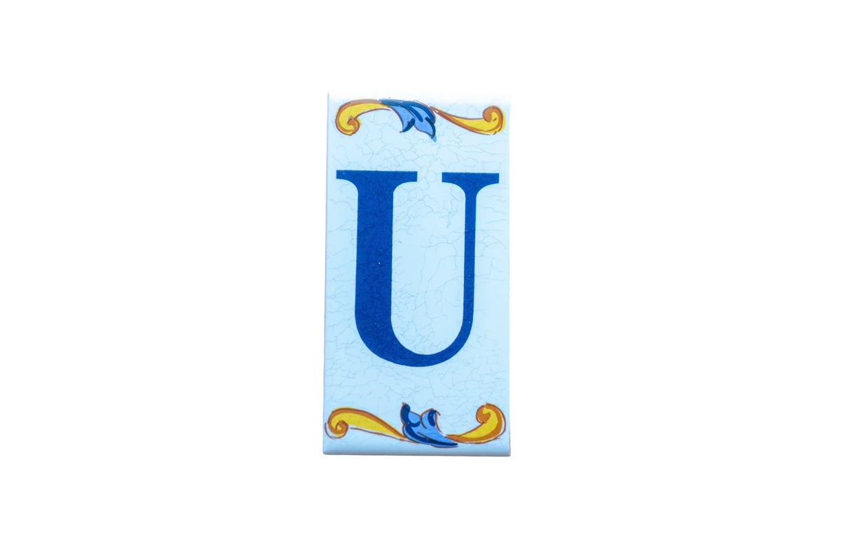 A Lisa Croquets Letters – Ceramic House Letter Tile featuring a blue letter U centered between decorative yellow and blue scrollwork on a light blue background.