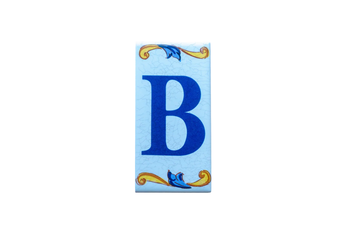 A Lisa Croquets Letters – Ceramic House Letter Tiles rectangular tile featuring a blue letter B centered between yellow and blue scrolls on a light blue crackle-finish background.