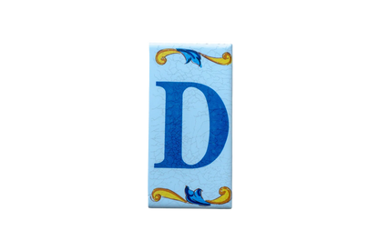A rectangular Lisa Croquets Letters – Ceramic House Letter Tile featuring the blue letter D centered between decorative yellow and blue scrollwork against a light blue, crackled-texture background.