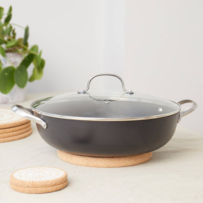 A black saucepan with a glass lid and metal handles rests on the LIGA Natural Cork Trivet - Round Small, protecting the light tablecloth. Coasters and a potted plant enhance the eco sustainable homeware feel in the background.