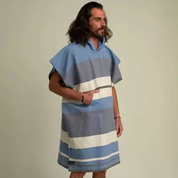 A man with long hair wears the Futah Papua Adult Beach Poncho—a premium Portuguese cotton surf poncho featuring blue, white, and gray stripes, short sleeves, and a front pocket—while standing against a plain background.