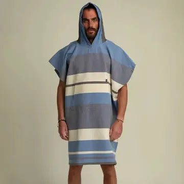 A man stands facing forward, wearing the Futah Papua Adult Beach Poncho—a hooded surf poncho made from premium Portuguese cotton, featuring wide blue, gray, and white horizontal stripes. The background is plain and neutral.