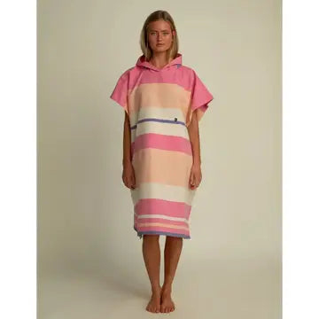 A woman stands barefoot against a plain, light background, wearing the Futah Papua Adult Beach Poncho—a loose, hooded surf poncho crafted from premium Portuguese cotton and featuring pink, white, and peach horizontal stripes.