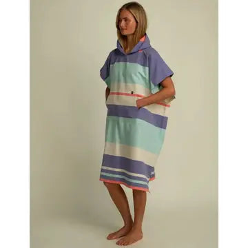 A woman stands barefoot wearing the Futah Papua Adult Beach Poncho, a hooded, short-sleeve surf poncho that falls to her knees and showcases wide pastel stripes in blue, green, cream, and coral. Made from premium Portuguese cotton, this beach poncho features a convenient front pocket where she rests her hands as she gazes thoughtfully to the side.