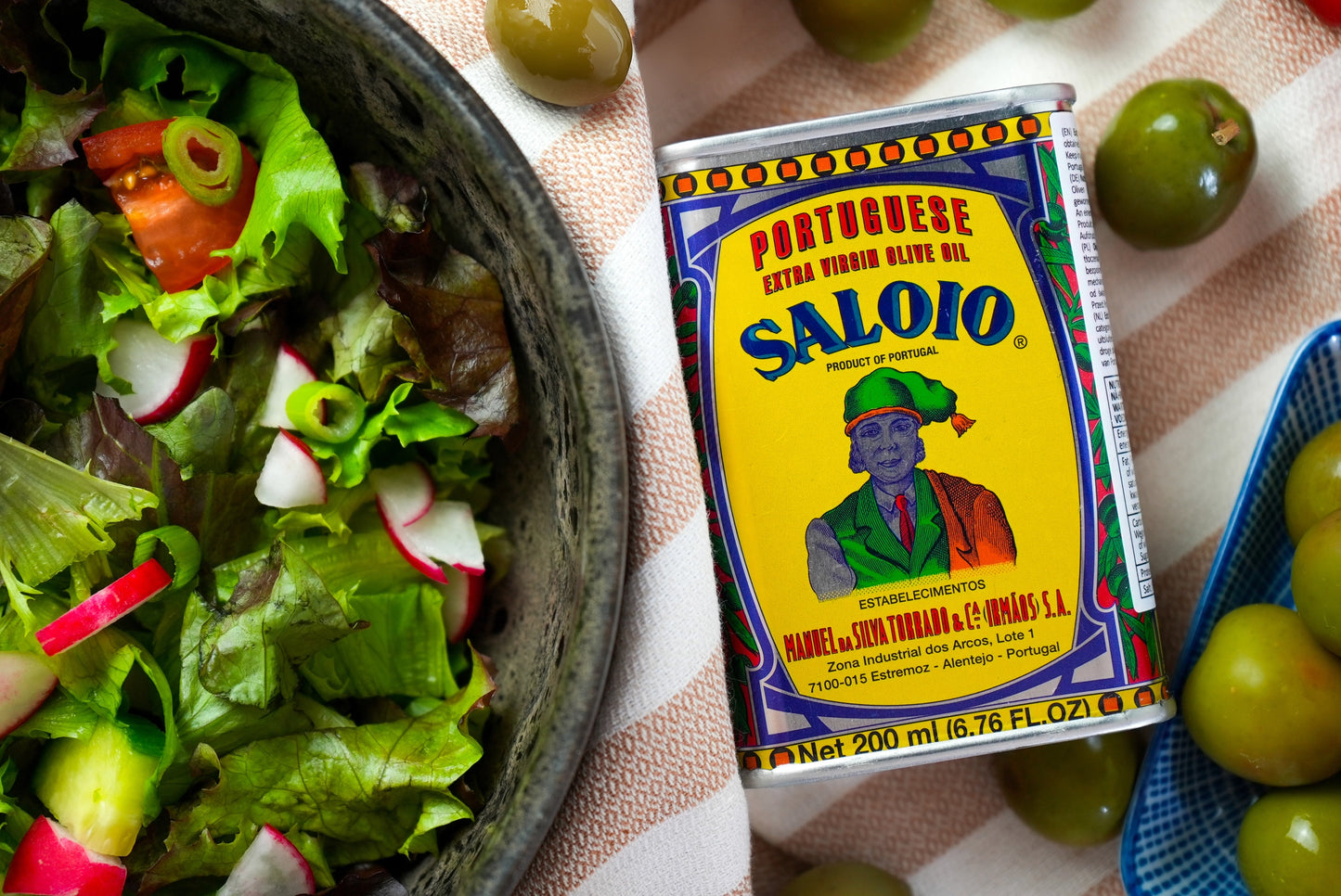 A can of Iberica's Sardine sampler kit #1 sits on a striped cloth next to salad, green olives, and a cheese board—perfect for an authentic Mediterranean spread with Pretty things from Portugal.