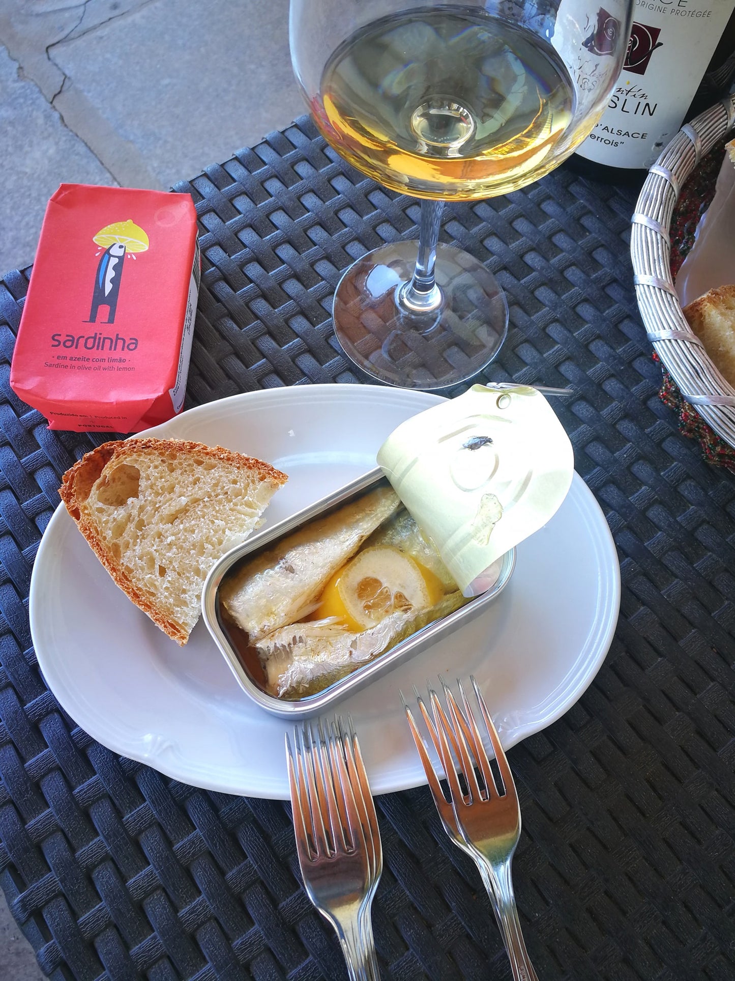 An open Sardinha Sardines in Lemon tin with fresh slices sits on a white plate next to bread. Two forks, the red tin, and a glass of white wine rest on the woven table—an omega-3 rich snack awaits.